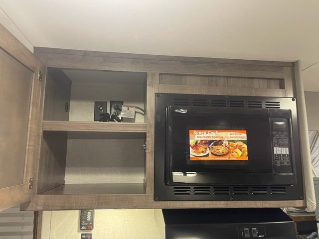 2-horse-gooseneck-trailer-with-living-quarters-microwave A kitchen cabinet with an open door showing an empty shelf and a power outlet inside, next to a black microwave with a recipe card displayed on its door. The setup is built into a wooden cabinet structure.