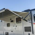 A silver RV with an extended black awning is parked outdoors under a clear blue sky. The door and windows are visible, along with part of a trailer attached next to it.