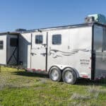 A silver horse trailer with a graphic of a running horse is parked on grass near a road and white fence, with clear skies and green hills in the background.