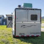 A silver horse trailer with SAFETACK REVERSE and a horse graphic parked on grass near a house, white fence, and hills under a clear sky.