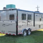 A large, shiny silver trailer with windows and horse graphics is parked on grass. The trailer has double axles, vents, and a green box on top, with SAFE-TACK REVERSE written on the back. A house and hills are in the background.