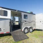 A silver horse trailer with two open doors and a ramp leads into the interior. The trailer is parked on grass, with white fencing and hills visible in the background under a clear blue sky.