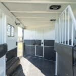 Interior of a clean, empty horse trailer with padded dividers, rubber flooring, and windows with metal bars, designed for safely transporting horses. Sunlight streams in through the open door and windows.