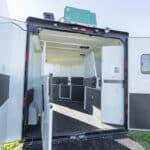 The image shows the open rear doors of a white horse trailer, revealing a clean, spacious interior with sections for transporting animals, non-slip flooring, vents, and storage areas under a bright, clear sky.
