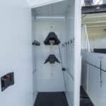 A white storage locker with open door showing hanging hooks, two black saddle racks mounted on the back wall, and a black rubber floor mat inside.