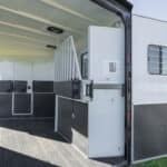 The image shows the interior of an empty horse trailer with open doors, padded dividers, rubber flooring, and windows with bars, parked in a rural outdoor setting.