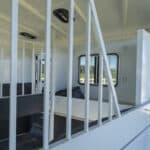 The interior of a horse trailer with white metal dividers, black padding, and windows letting in natural light. The door is open, showing a glimpse of the outdoors and a wooden fence.