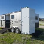A silver horse trailer with two ramps down and a slide-out section is parked on grass near a white fence under a clear blue sky.