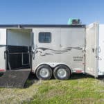 A silver horse trailer with two doors open, one showing a ramp and the other showing storage space. The trailer is parked on grass under a clear blue sky. Black decorative lines are on the side.