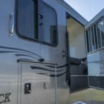 A close-up of a gray trailer with a door and window open, showing part of the interior. Stylized black graphics decorate the exterior. Bright daylight and a clear blue sky are visible.