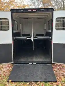 Open double doors reveal the interior of an empty horse trailer with padded dividers, rubber flooring, and a lowered ramp, surrounded by fallen autumn leaves outside.