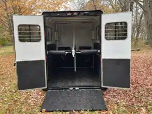 Open horse trailer with double rear doors and ramp down, showing a clean, empty interior with padded dividers. The trailer is parked on fallen autumn leaves with trees in the background.