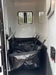 The image shows the interior of a horse trailer with two large black feed bags secured by straps below a window with metal bars. The area is clean and organized, with a white divider on the left side.