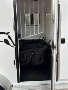 The image shows the open side door of a horse trailer, revealing the interior with black feed bags hanging on partitions and a barred window at the front. The trailer is white with a partial horse graphic on the outside.