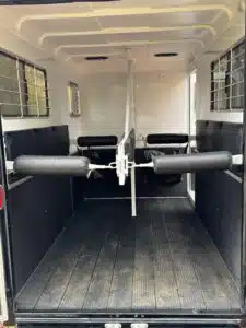 The interior of a horse trailer with padded dividers, black rubber flooring, white walls, and metal bars on the windows, designed for safely transporting two horses.