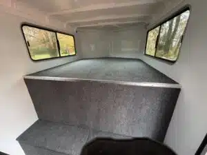 Interior of a vehicle or camper with carpeted raised sleeping or storage platform, large windows on both sides, and trees visible outside through the windows.
