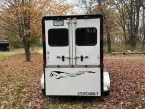 A white horse trailer with two rear doors, black trim, and the word SPORTABOUT below a running horse graphic, parked on grass among autumn trees.