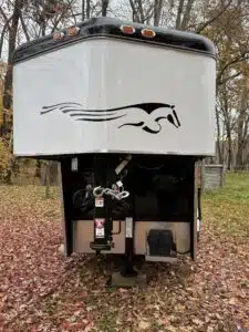 A white and black horse trailer with a stylized running horse decal on the front, parked on fallen autumn leaves with trees in the background.